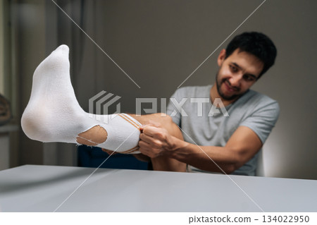 Unlucky young man sitting at table, tearing white sock on leg, while trying to put it on foot, revealing hole near ankle, illustrating resourcefulness, financial hardship, need for new garments. Unlucky young man sitting at table, tearing white sock on leg, while trying to put it on foot, revealing hole near ankle, illustrating resourcefulness, financial hardship, need for new garments. 134022950