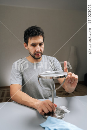 Vertical portrait of focused bearded man cleaning small desktop vanity mirror with finger, examining surface for smudges and dust, focusing on detailed household chores and personal hygiene routine. 134023001