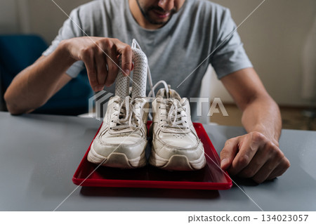 Closeup cropped shot of young man inserting portable electric dryer into white sneakers on red tray, demonstrating footwear care, moisture removal and hygienic drying to prevent odor and damage. Closeup cropped shot of young man inserting portable electric dryer into white sneakers on red tray, demonstrating footwear care, moisture removal and hygienic drying to prevent odor and damage. 134023057