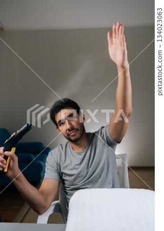 Vertical shot of curious man feeling bored with everyday chores, playfully engaging with steam cleaner, shooting vapor and raising hand, distracting himself from household cleaning routine. 134023063