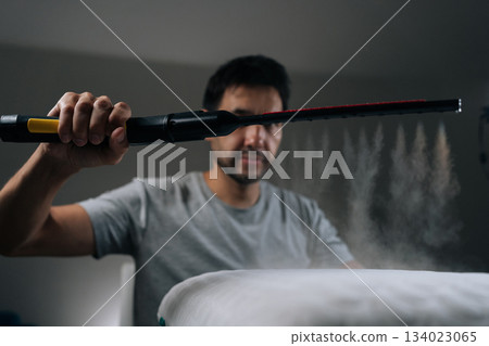 Front view man using handheld steam sterilizer tool for deep cleaning and disinfecting white memory foam pillow, ensuring hygiene and eliminating allergens from bedding. Concept of household chores. 134023065