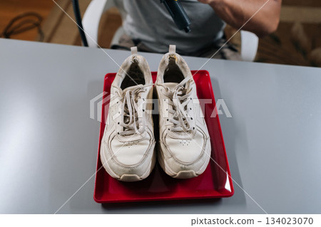 Person preparing to steam-cleans and dries dirty white sneakers on red tray, restoring bright, fresh appearance while demonstrating careful at home shoe care and stain removal routine. 134023070