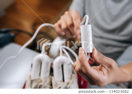Person plugs portable electric shoe dryer into power to dry and sanitize white sneakers, keeping footwear fresh, hygienic and ready for daily wear with modern home tech, close-up. 134023082