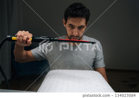 Man holding using steam cleaner tool over white memory foam pillow, sending vapor onto fabric to sanitize and deep clean bedding item, representing household care and allergy control. 134023084