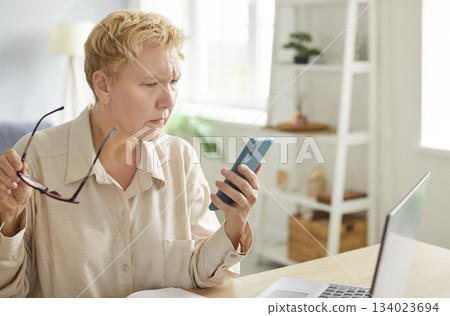 Focused woman with blond hair looking at smartphone screen while sitting at desk on workplace. Focused woman with blond hair looking at smartphone screen while sitting at desk on workplace. 134023694