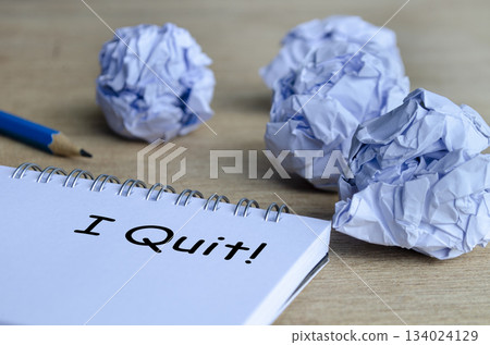 A dramatic workspace scene with crumpled paper surrounding a notepad that displays the handwritten message I Quit, symbolizing stress and burnout. 134024129