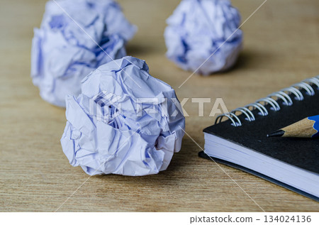 A closeup view of crumpled sheets of paper resting near a spiral notebook and pencil. Work in progress concept. A closeup view of crumpled sheets of paper resting near a spiral notebook and pencil. Work in progress concept. 134024136