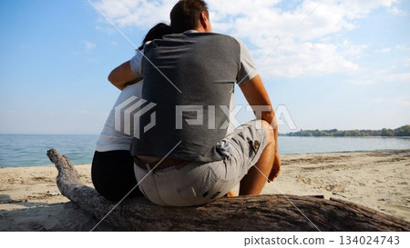 Happy pair hugs during sitting on wooden log on beach. Romantic couple admiring sea landscape at sunny day. Lovers spending time together at resort. Concept of summer vacation or holiday. Slow mo 134024743