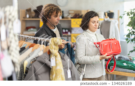 Fashionable female client standing against background of shelves with clothes choosing handbag and male salesperson or husband helping with assortment and choice in clothing store Fashionable female client standing against background of shelves with clothes choosing handbag and male salesperson or husband helping with assortment and choice in clothing store 134026189