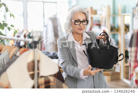 Mature woman standing in store and choosing stylish bag Mature woman standing in store and choosing stylish bag 134026249