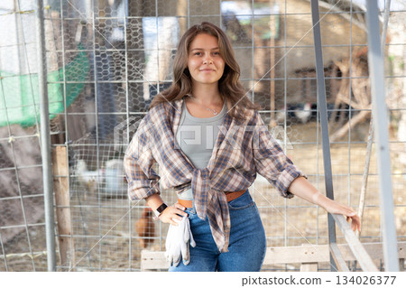 Young woman owner of livestock farm stands near corral mesh fence for livestock and rests Young woman owner of livestock farm stands near corral mesh fence for livestock and rests 134026377