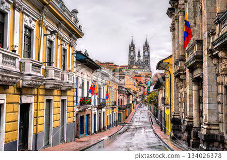 Cobblestone street leads toward Basilica del Voto Nacional in Quito, Ecuador. Historic center landscape features colonial buildings and wet road under cloudy sky in UNESCO World Heritage site 134026378