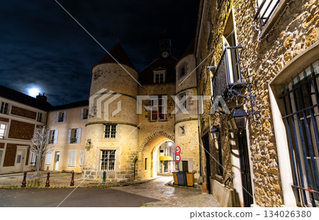 Night view of the historic Gate of Garlande Castle and the town hall in Tournan-en-Brie, Paris Region of France. The medieval stone gatehouse with a clock tower is illuminated under a dark sky 134026380