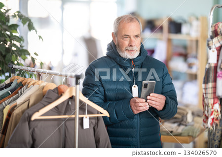 Senior man is fitting on and trying on down jacket in store and take pictures in new clothes, wants to capture new look. Buyer looks at himself in mirror while shopping, evaluates appearance. Senior man is fitting on and trying on down jacket in store and take pictures in new clothes, wants to capture new look. Buyer looks at himself in mirror while shopping, evaluates appearance. 134026570