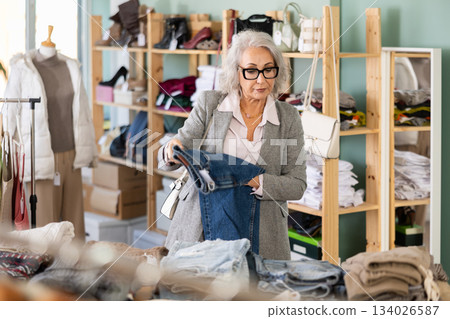 Mature female buyer in glasses chooses jeans in clothing store 134026587