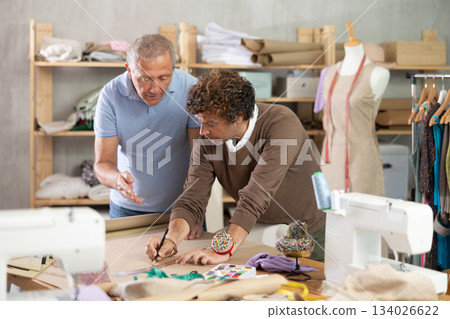 Elderly man tailor teaches adult man to draw pattern on paper Elderly man tailor teaches adult man to draw pattern on paper 134026622