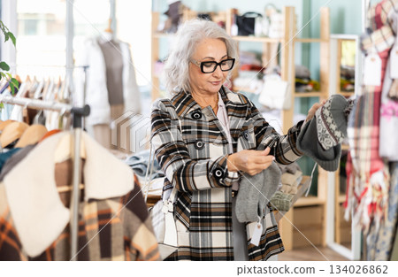 Mature woman in glasses chooses fashionable warm hat in clothing store 134026862