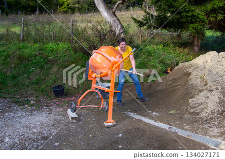Woman puts cement with a shovel into the cement mixer. Woman puts cement with a shovel into the cement mixer. 134027171