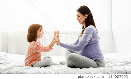 Adorable little girl playing with her pregnant mom, sitting on bed at home, mother and daughter touching palms, family connection concept Adorable little girl playing with her pregnant mom, sitting on bed at home, mother and daughter touching palms, family connection concept 134027493