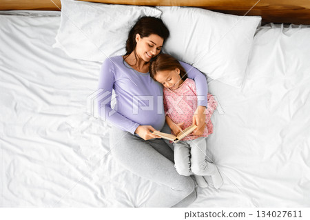 Top view of loving pregnant mom reading book to her little daughter while resting together in bed, top view Top view of loving pregnant mom reading book to her little daughter while resting together in bed, top view 134027611