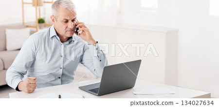 Business Calls Concept. Concerned Senior Man Talking On Mobile Phone Sitting At Desk, Looking At Laptop, Empty Space Business Calls Concept. Concerned Senior Man Talking On Mobile Phone Sitting At Desk, Looking At Laptop, Empty Space 134027634