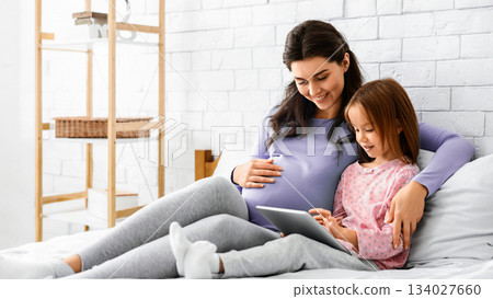 Beautiful pregnant mother and her cute little daughter using digital tablet and smiling while spending time together in bed 134027660