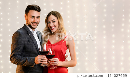 Young Elegant Couple Posing With Red Wine Glasses In Hands, Standing Over Beige Background And Looking At Camera, Free Space Young Elegant Couple Posing With Red Wine Glasses In Hands, Standing Over Beige Background And Looking At Camera, Free Space 134027669