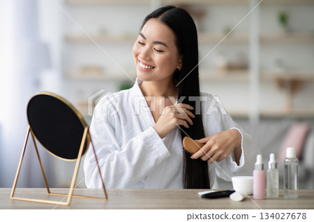 Cheerful young asian lady in bathrobe sitting in bedroom, combing her beautiful silky hair with wooden brush, looking at mirror and smiling, copy space. Hair care products concept Cheerful young asian lady in bathrobe sitting in bedroom, combing her beautiful silky hair with wooden brush, looking at mirror and smiling, copy space. Hair care products concept 134027678