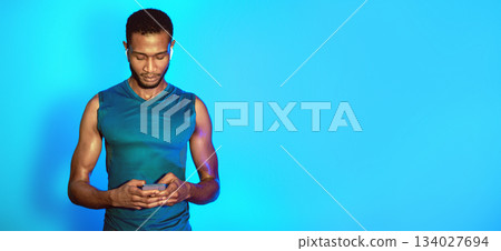 Sporty Black Man Using Phone And Earbuds Earphones Standing On Blue Studio Background. Playlist For Fitness. Copy Space Sporty Black Man Using Phone And Earbuds Earphones Standing On Blue Studio Background. Playlist For Fitness. Copy Space 134027694