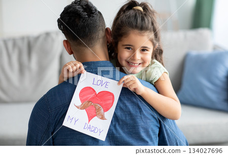 Father's Day Concept. Smiling little girl embracing dad and holding greeting card with drawn big red heart, greeting daddy with holiday, cheerful female child looking at camera, closeup shot Father's Day Concept. Smiling little girl embracing dad and holding greeting card with drawn big red heart, greeting daddy with holiday, cheerful female child looking at camera, closeup shot 134027696
