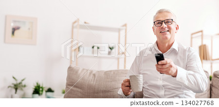Watching Movie On Tv. Smiling Senior Man Sitting On Couch With Remote Controller, Holding Coffee Mug, Copyspace Watching Movie On Tv. Smiling Senior Man Sitting On Couch With Remote Controller, Holding Coffee Mug, Copyspace 134027768