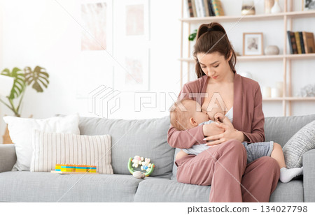 Breastfeeding Concept. Beautiful young woman lactating her toddler baby on couch at home, copy space 134027798
