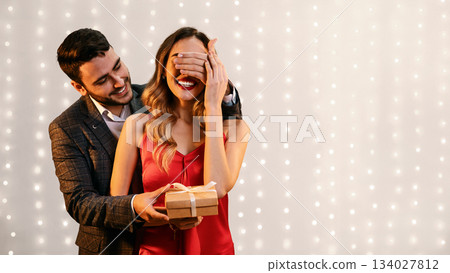 Romantic Surprise. Elegant man covering his girlfriend's eyes and giving her present for Valentine's Day or Birthday, standing together indoors 134027812