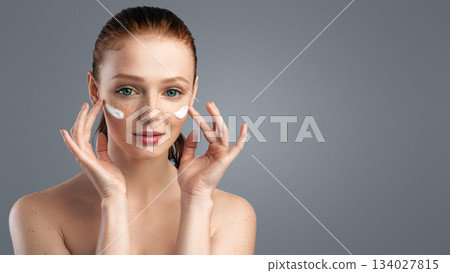 Young Female Applying Face Cream On Cheeks With Both Hands Caring For Facial Skin Looking At Camera Over Gray Background. Studio Shot. Skin Care Cosmetics 134027815