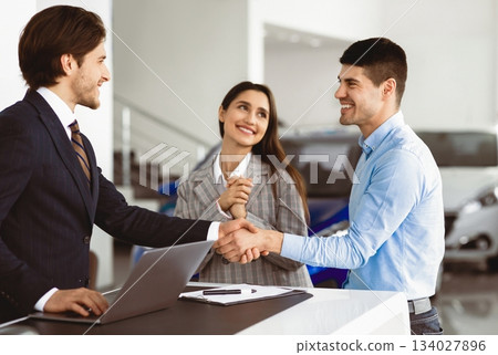 Young Spouses Buying Car, Husband Handshaking With Seller Man Standing In Dealership Office. Selective Focus Young Spouses Buying Car, Husband Handshaking With Seller Man Standing In Dealership Office. Selective Focus 134027896