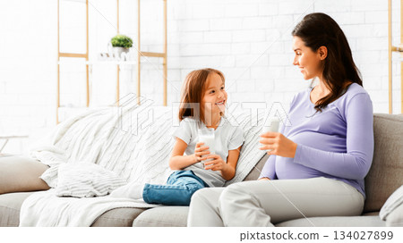 Pregnant loving mother drinking milk with her smiling little daughter, sitting on sofa at living room Pregnant loving mother drinking milk with her smiling little daughter, sitting on sofa at living room 134027899