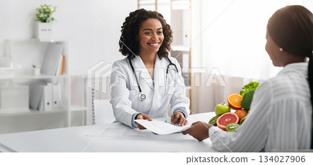 Personal diet. Cheerful african american female dietologist giving diet plan to woman client, clinic interior 134027906