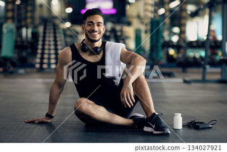Young Arab Male Athlete Relaxing On Floor After Training At Gym, Smiling Middle Eastern Man Resting Next To Blank Container With Supplement Pills, Enjoying Bodybuilding And Healthy Lifestyle 134027921