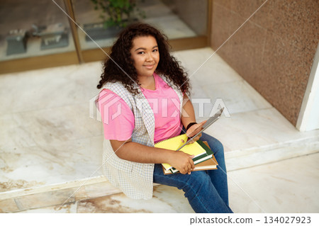 A young student sits on the stairs, casually studying with a tablet and a stack of notebooks. She appears focused and engaged in her work, surrounded by a warm atmosphere. A young student sits on the stairs, casually studying with a tablet and a stack of notebooks. She appears focused and engaged in her work, surrounded by a warm atmosphere. 134027923