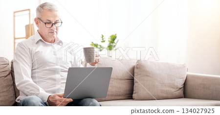 Reading Morning News. Focused Mature Man In Specs Using Laptop And Drinking Hot Tea, Sitting On Couch, Copyspace Reading Morning News. Focused Mature Man In Specs Using Laptop And Drinking Hot Tea, Sitting On Couch, Copyspace 134027925