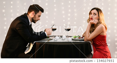 Failed Date. Unhappy young woman sitting alone while her boyfriend stuck into his phone during dinner in restaurant, side view with free space Failed Date. Unhappy young woman sitting alone while her boyfriend stuck into his phone during dinner in restaurant, side view with free space 134027931
