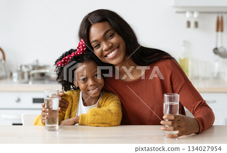 Happy African American Family Mother And Daughter Holding Glasses With Water While Sitting Together At Table In Kitchen, Smiling Black Mom And Female Child Enjoying Healthy Drink, Free Space 134027954