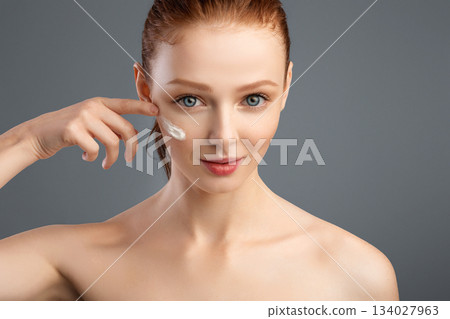Skincare Routine. Young Attractive Female Applying Facial Moisturizing Cream On Cheek Posing Standing Shirtless In Studio Over Gray Background. Facial Skin Care And Beauty. Cropped Skincare Routine. Young Attractive Female Applying Facial Moisturizing Cream On Cheek Posing Standing Shirtless In Studio Over Gray Background. Facial Skin Care And Beauty. Cropped 134027963