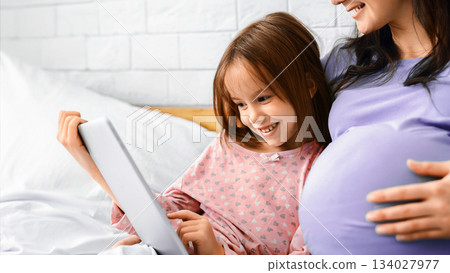 Close up of happy little girl using digital tablet with pregnant mom, home interior 134027977