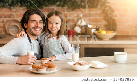 Healthy Snacks. Portrait of happy little kid girl and her dad eating muffins, drinking milk and posing to camera in kitchen Healthy Snacks. Portrait of happy little kid girl and her dad eating muffins, drinking milk and posing to camera in kitchen 134027989