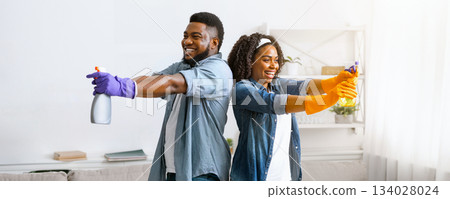 On guard of cleanliness. Cheerful black couple having fun while cleaning apartment, aiming with detergent sprayers, standing back to back and laughing 134028024