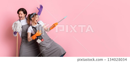 Young couple of janitors having fun during cleaning, fooling with mop and broom over pink background, panorama with copy space 134028034