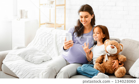 Smiling pregnant woman taking selfie with her little daughter while sitting on sofa at home, free space 134028060