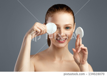 Joyful Young Female Covering Eye With Cotton Pad Posing And Having Fun Caring For Face Skin Standing On Gray Background, Smiling Looking At Camera. Studio Shot. Facial Skincare Concept 134028070
