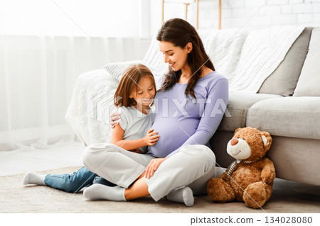 Family concept. Adorable little girl cuddling with her expecting mother at home, sitting on floor and playing with teddy bear, copy space Family concept. Adorable little girl cuddling with her expecting mother at home, sitting on floor and playing with teddy bear, copy space 134028080